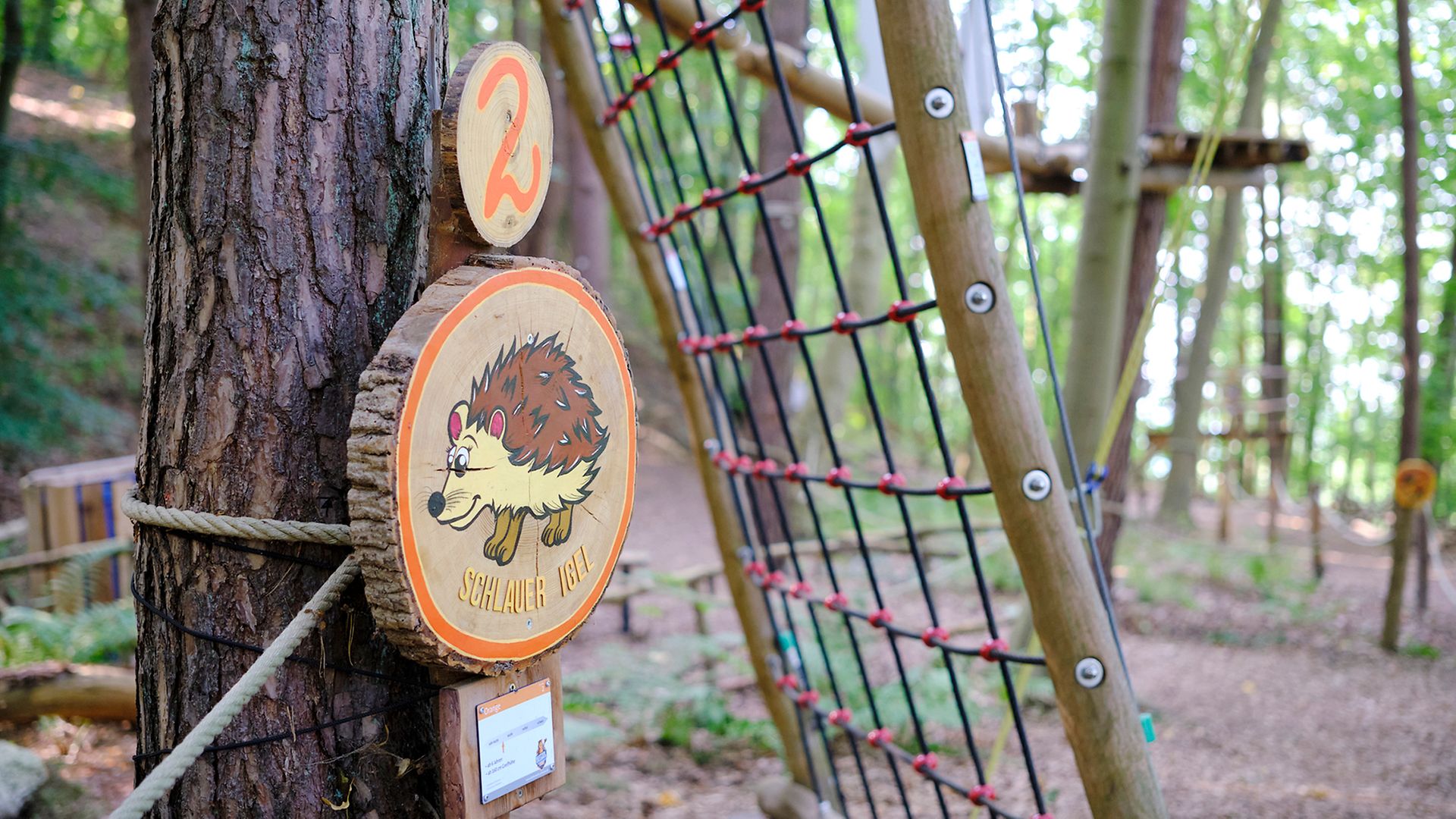 Im Vordergrund ist ein Holzschild zu sehen, auf dem "Schlauer Igel" und eine "2" stehen, im Hintergrund der Kletterpark.