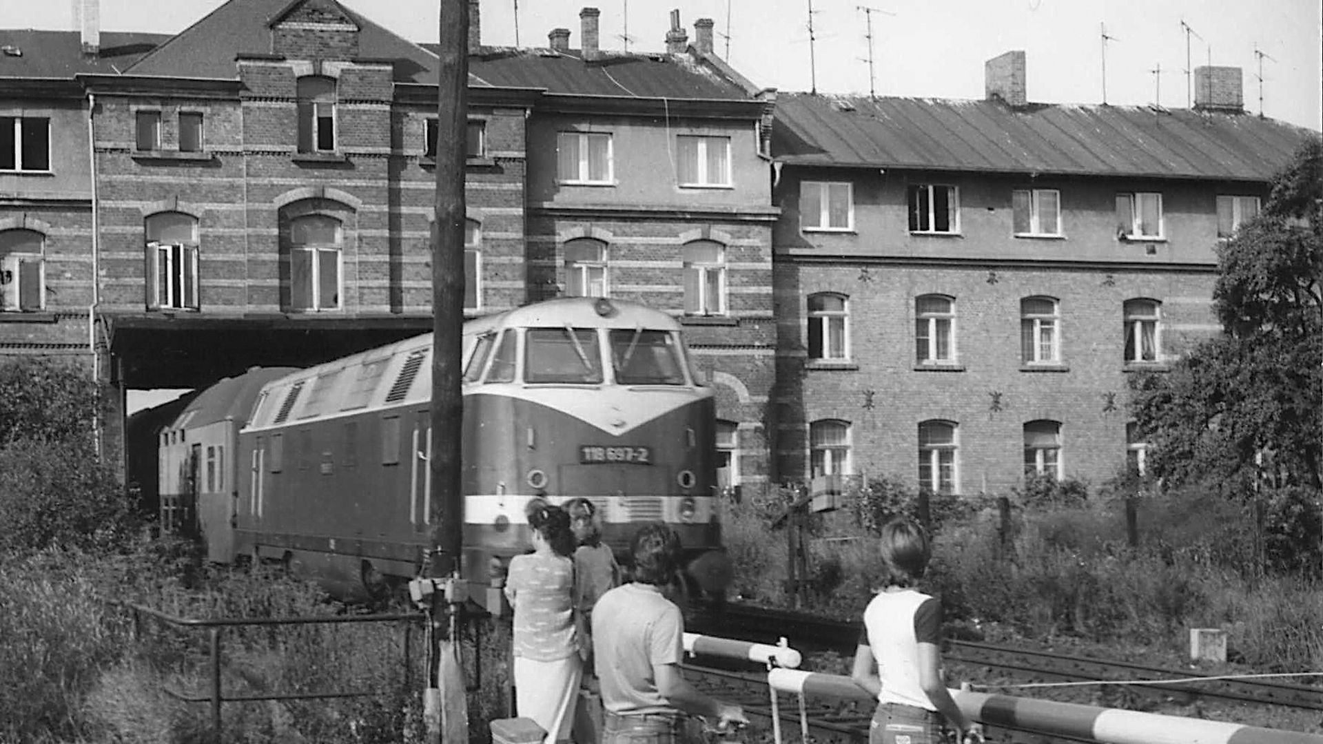 Schwarzweiß-Bild: Eine S-Bahn fährt durch einen Tunnel, der durch ein Haus führt. An einer Schranke warten Fahrradfahrende.