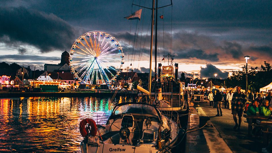 Blick auf Wolgaster Hafen bei Abendstimmung mit beleuchtetem Riesenrad und Marktständen.