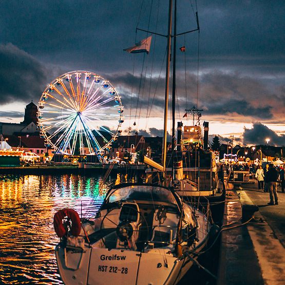 Blick auf Wolgaster Hafen bei Abendstimmung mit beleuchtetem Riesenrad und Marktständen.