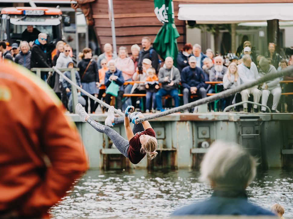 Eine Wettkampfteilnehmerin hangelt sich vor zahlreichen Schaulustigen an einem Seil über den Fluss Ryck in Greifswald