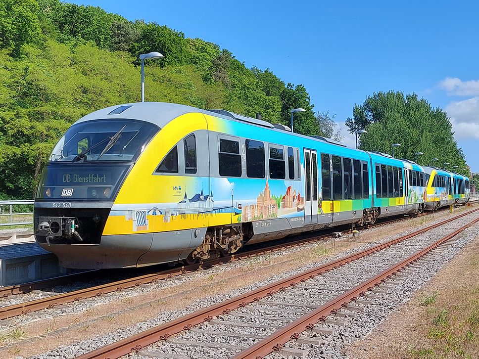 Ein mit MV-Motiven folierter Regionalzug des Typs BR 642 steht am Bahnhof vor einer Baumgruppe