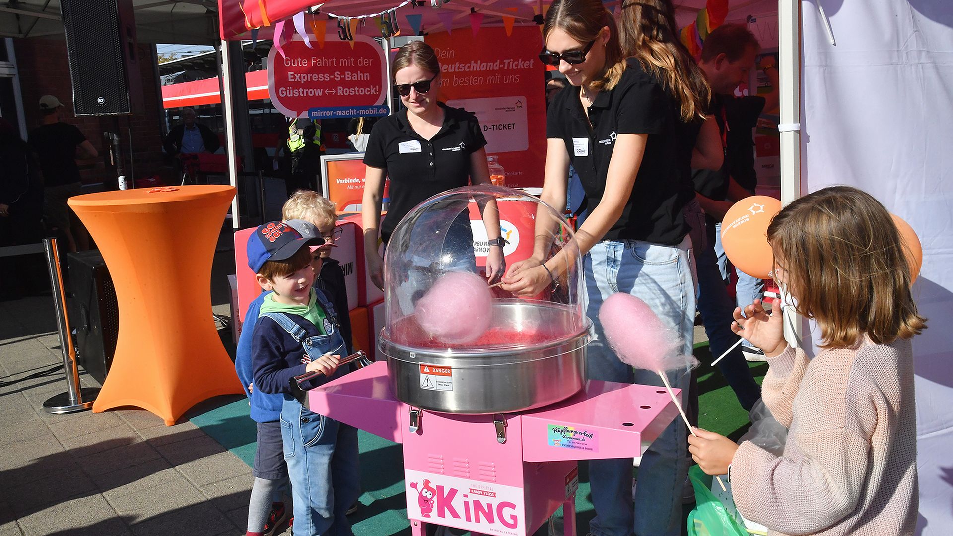 Zwei Kinder stehen an einem Stand, an dem rosa Zuckerwatte verteilt wird.