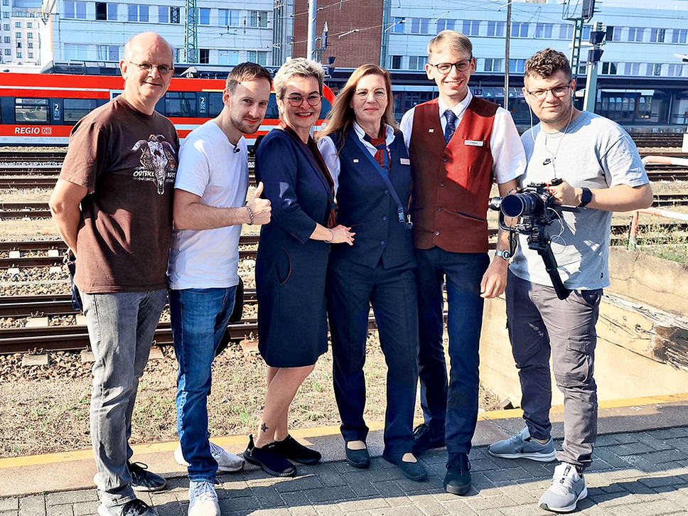 Zwei Männer, zwei Frauen in Bahn-Uniform, ein Kundenbetreuer und ein Mann mit einer Kamera am Bahnsteig vor einem Regionalzug.