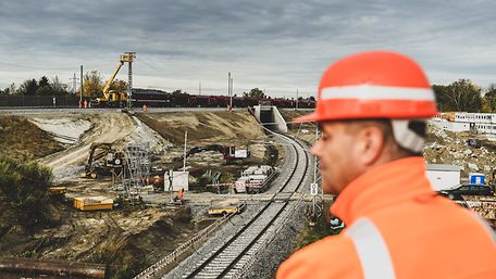 Mann mit Bauhelm in Nahaufnahme vor Baustelle an Bahngleisen